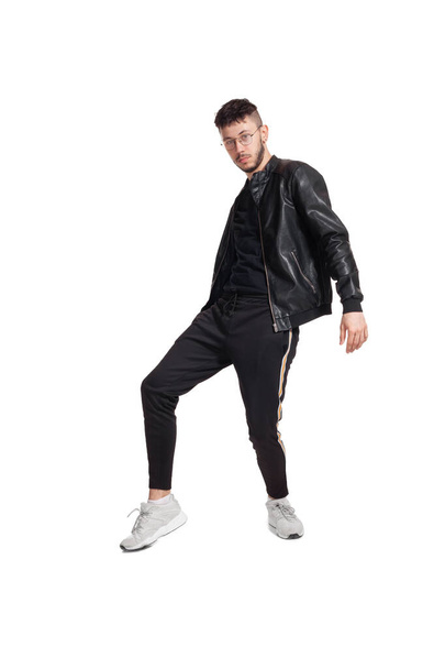 Full-length photo of a modern performer in glasses, black leather jacket, t-shirt, sports pants and light sneakers fooling around in studio. Indoor photo of an athletic guy dancing and showing his leg - Foto, afbeelding