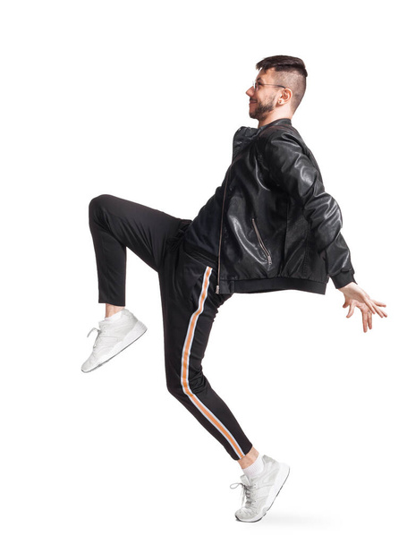 Full-length photo of a modern performer in glasses, black leather jacket, t-shirt, sports pants and light sneakers fooling around sideways in studio. Indoor photo of an athletic man dancing and - Foto, imagen