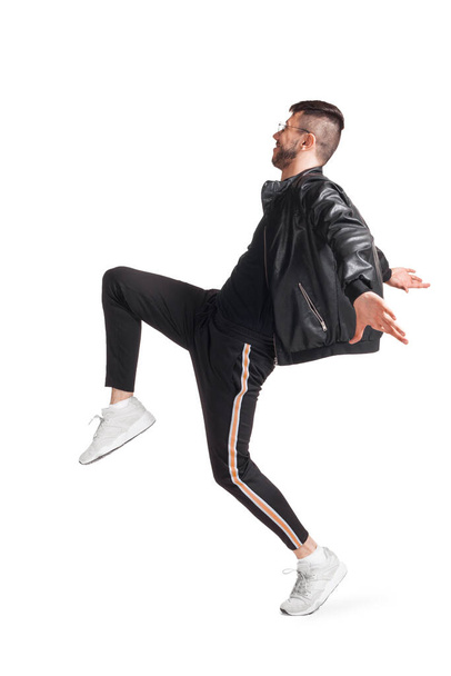 Full-length photo of a modern performer in glasses, black leather jacket, t-shirt, sports pants and light sneakers fooling around in studio. Indoor photo of a nice person dancing sideways isolated on - Foto, immagini