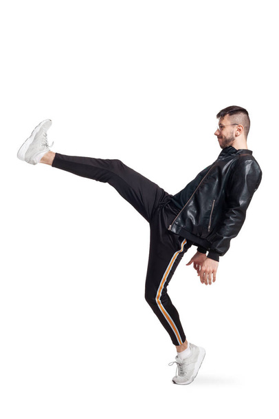 Full-length photo of a modern performer in glasses, black leather jacket, t-shirt, sports pants and light sneakers fooling around in studio. Indoor photo of a nice fellow dancing and raising his leg - Φωτογραφία, εικόνα