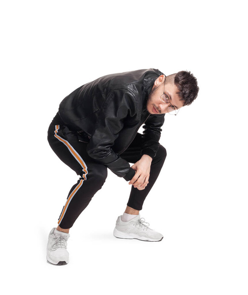 Full-length photo of a modern performer in glasses, black leather jacket, t-shirt, sports pants and light sneakers fooling around in studio. Indoor photo of a nice man dancing and looking at the - Fotó, kép
