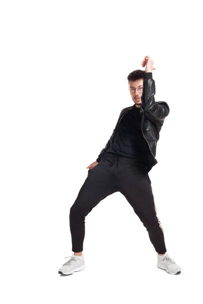 Full-length photo of a modern performer in glasses, black leather jacket, t-shirt, sports pants and light sneakers fooling around in studio. Indoor photo of an attractive person dancing isolated on - Foto, immagini