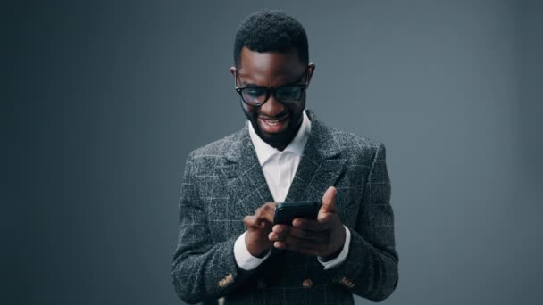 Afrikai-amerikai üzletember kezében egy telefont néz rá, és boldog, hogy nyerni és nyerni az irodában szürke alapon. Kiváló minőségű 4k felvételek - Felvétel, videó