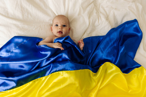 Portrait of a baby wrapped in the national blue and yellow flag of Ukraine, lying on the bed. Baby with Ukraine flag heart. photo with place for text. fear and expectation. children's tears - 写真・画像