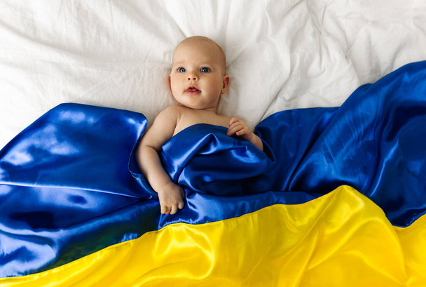 Portrait of a baby wrapped in the national blue and yellow flag of Ukraine, lying on the bed. Baby with Ukraine flag heart. photo with place for text. Children of war. The child is scared - Photo, Image