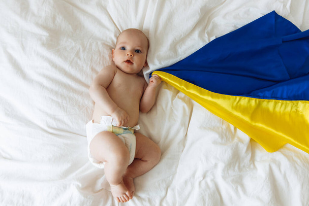 Portrait of a baby wrapped in the national blue and yellow flag of Ukraine, lying on the bed. Baby with Ukraine flag heart. photo with place for text. Children of war - Valokuva, kuva