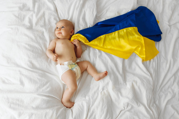 Portrait of a baby wrapped in the national blue and yellow flag of Ukraine, lying on the bed. Baby with Ukraine flag heart. photo with place for text. Children of war - Fotografie, Obrázek