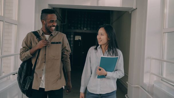 Young attractive african american male and asian female students are moving down corridor discussing something lively - Foto, Imagem
