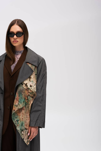 teenage model in stylish sunglasses and trench coat isolated on grey - Zdjęcie, obraz