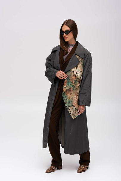 full length of brunette teenage model in stylish sunglasses and trendy trench coat on grey - Foto, afbeelding