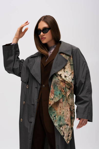 stylish teenage model in sunglasses and trendy trench coat posing isolated on grey - Foto, Imagem