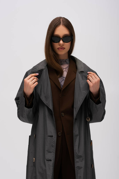stylish teenage girl in sunglasses and trendy trench coat posing isolated on grey - Fotó, kép