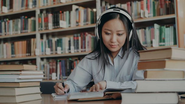Pretty positive smiling asian student with headphones on head listening to music is sitting at table in university library holding book and writing down summary - Photo, Image