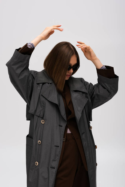 teenage model in sunglasses and trendy trench coat posing isolated on grey - Foto, imagen