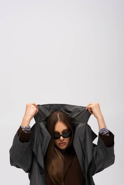 stylish teenage model in sunglasses adjusting collar of trench coat isolated on grey - 写真・画像