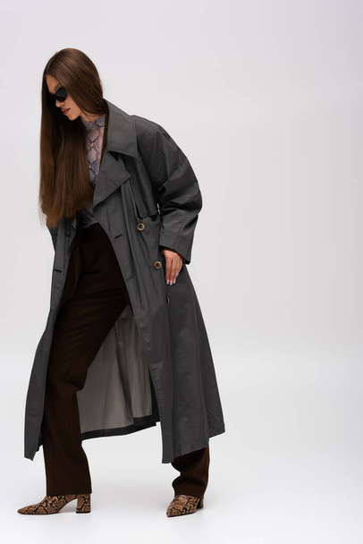 full length of teenage model in sunglasses and trench coat posing on gray - Photo, Image