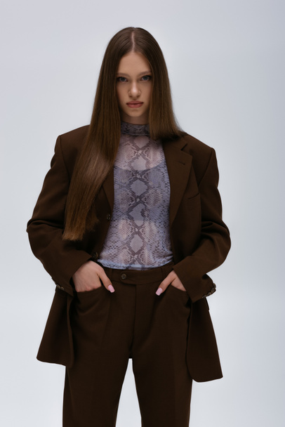 pretty teenage model in brown high-quality suit posing with hands in pockets isolated on grey - Fotografie, Obrázek