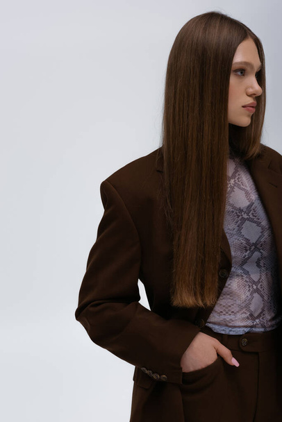 pretty teenage model in brown high-quality suit posing with hand in pocket isolated on grey - Foto, afbeelding