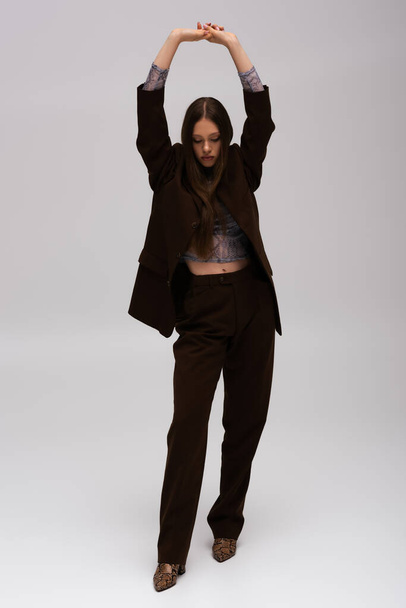 full length of teenage model in stylish high-quality suit posing with raised hands on grey - Foto, Imagen