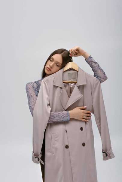 brunette teenage model holding recyclable trench coat on hanger isolated on grey - Foto, imagen
