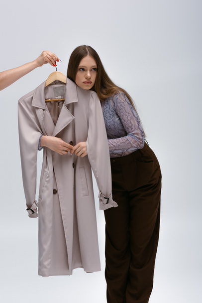 teenage model hugging recyclable trench coat while woman holding hanger on grey - Fotoğraf, Görsel