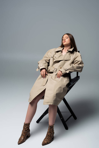 full length of model with closed eyes in stylish trench coat posing on chair of grey - Foto, imagen