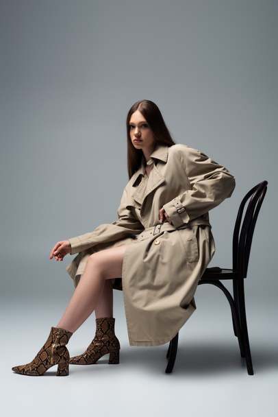 full length of brunette model in trench coat sitting on chair and posing on grey - Photo, Image