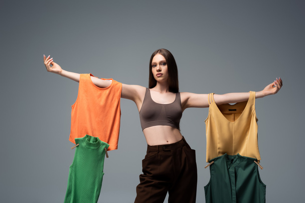 pretty model in crop top and trousers standing with outstretched hands and holding clothing on grey  - Fotoğraf, Görsel