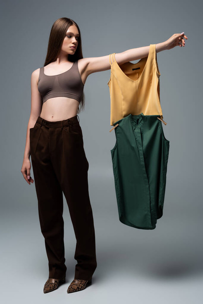 full length of brunette model in crop top and trousers standing with outstretched hand and holding clothing on grey - Fotoğraf, Görsel