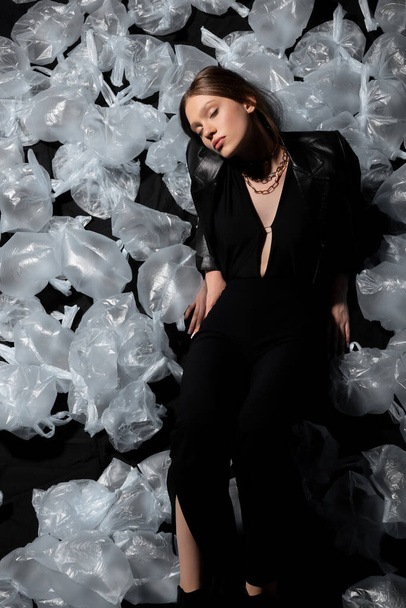top view of brunette model with closed eyes in stylish suit posing around plastic waste on black  - Photo, Image