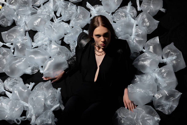 top view of brunette model in stylish suit posing around plastic bags on black  - Foto, afbeelding