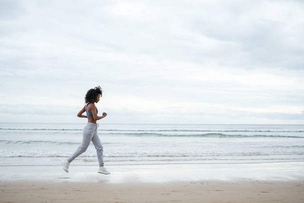 Sporty fit afro hairstyle woman running at the beach on a cloudy day. - Foto, immagini