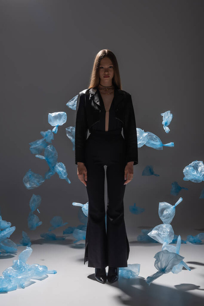 full length of pretty model in black suit posing near blue plastic bags on grey - Zdjęcie, obraz
