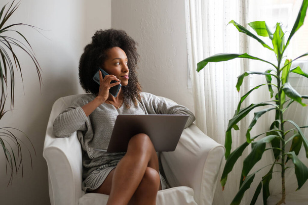 Black woman at home using laptop for working from home while doing a phone call. - Φωτογραφία, εικόνα