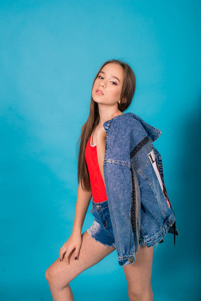 A cute teenage girl with long hair in trendy denim youth clothes and a red T-shirt on a blue background in the studio. Teenage fashion - Valokuva, kuva