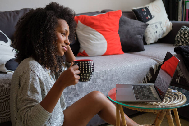 Comfortable relaxed casual young black woman drinking coffee and using laptop in living room at home. - Φωτογραφία, εικόνα