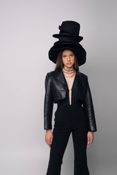 teenage model in leather jacket and different black hats on head isolated on grey - Foto, Imagem