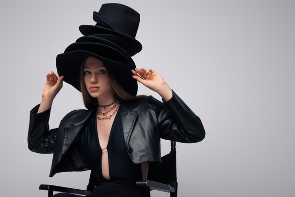 teenage model in leather jacket and different black hats sitting isolated on grey - Fotografie, Obrázek