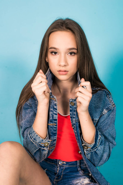 A cute teenage girl with long hair in trendy denim youth clothes and a red T-shirt on a blue background in the studio. Teenage fashion - Foto, imagen
