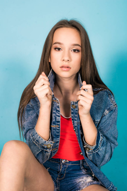 A cute teenage girl with long hair in trendy denim youth clothes and a red T-shirt on a blue background in the studio. Teenage fashion - Фото, зображення