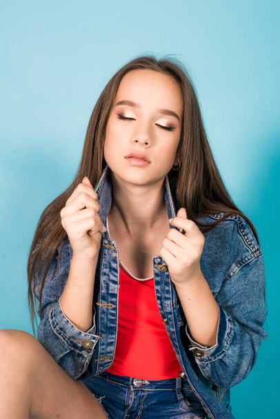 A cute teenage girl with long hair in trendy denim youth clothes and a red T-shirt on a blue background in the studio. Teenage fashion - Foto, Imagem