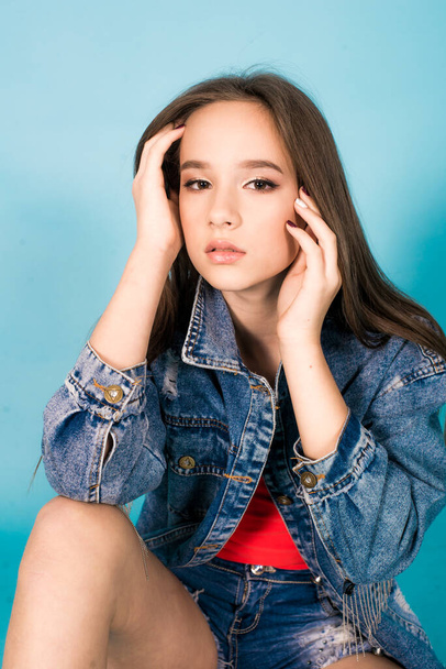 A cute teenage girl with long hair in trendy denim youth clothes and a red T-shirt on a blue background in the studio. Teenage fashion - Fotó, kép