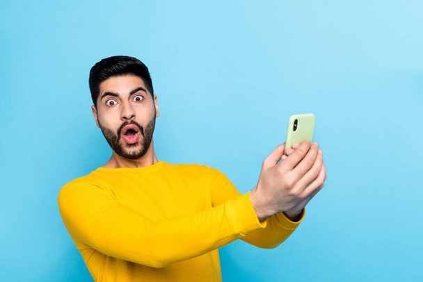 Photo of funky shocked man wear yellow sweater communicating modern device empty space isolated blue color background. - Zdjęcie, obraz