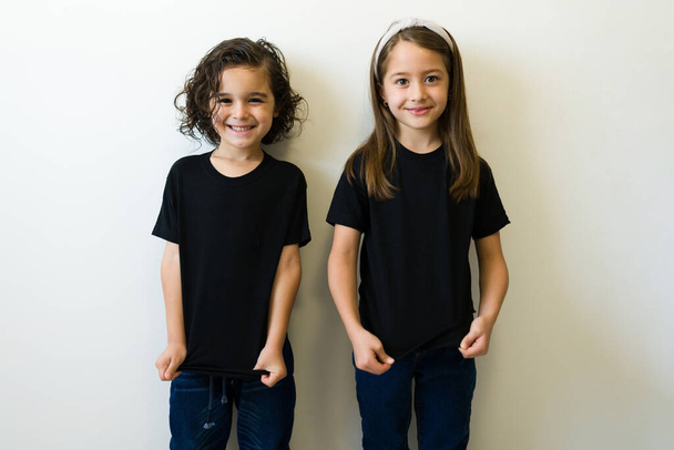 Cheerful boy and girl showing the front design print of their black mockup t-shirt against a white studio background - Foto, Bild