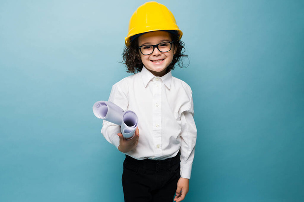 Happy little boy with glasses and a yellow helmet using construction plans while playing architect against a blue background - Foto, Imagen