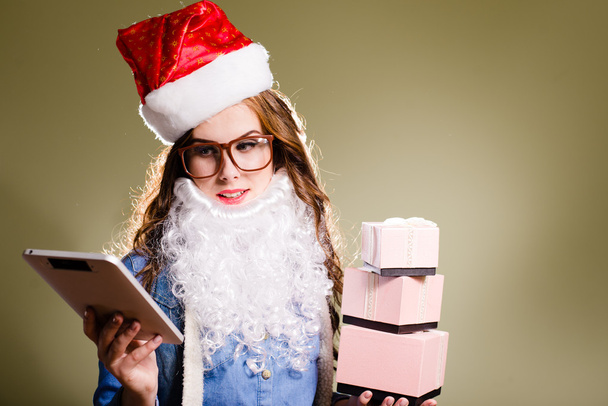 Funny hipster girl in super size eyeglasses wearing xmas santa hat and faux beard holding three boxes of presents and tablet pc over olive copy space background - Zdjęcie, obraz