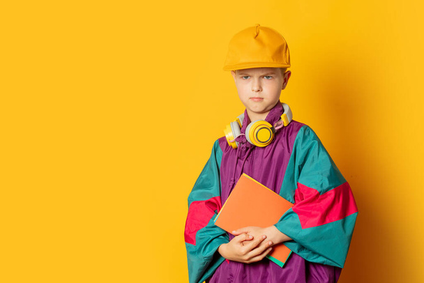 Stylish kid boy in 80s sport suit and cap with notebooks on yellow background - Fotó, kép