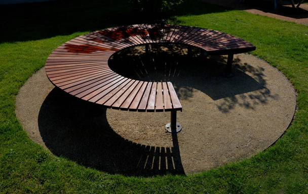 circular bench made of brown-red teak wood. tropical impregnated podia boards for sunbathing visitors in the park. spiral shape, snail shells. trowel base in the lawn, tree, trunk - Zdjęcie, obraz