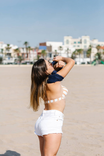 a pretty girl in a bikini taking off her sweater over her head on the beach with the city in the background - Foto, immagini