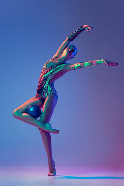Портрет молодої красивої дівчини, ритмічної гімнастки, яка позує з синім м'ячем ізольовано на синьому фіолетовому фоні в неоновому світлі. Концепція дії, руху, спортивного життя, мотивації, конкуренції
. - Фото, зображення